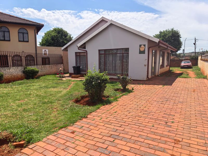 Front view of a detached house featuring a brick paved driveway, landscaped garden, and secure window bars, set within a fenced residential property.