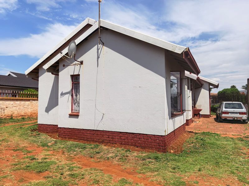 Rear view of the house featuring a brick plinth, metal window frames with security bars, and an external wash basin on a paved area.