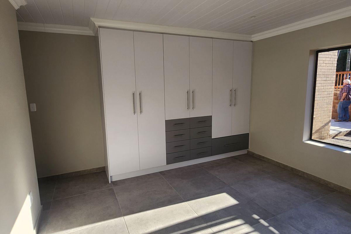 A bright bedroom featuring modern built-in cupboards with a two-tone finish, large format floor tiles, and a window overlooking the exterior area.