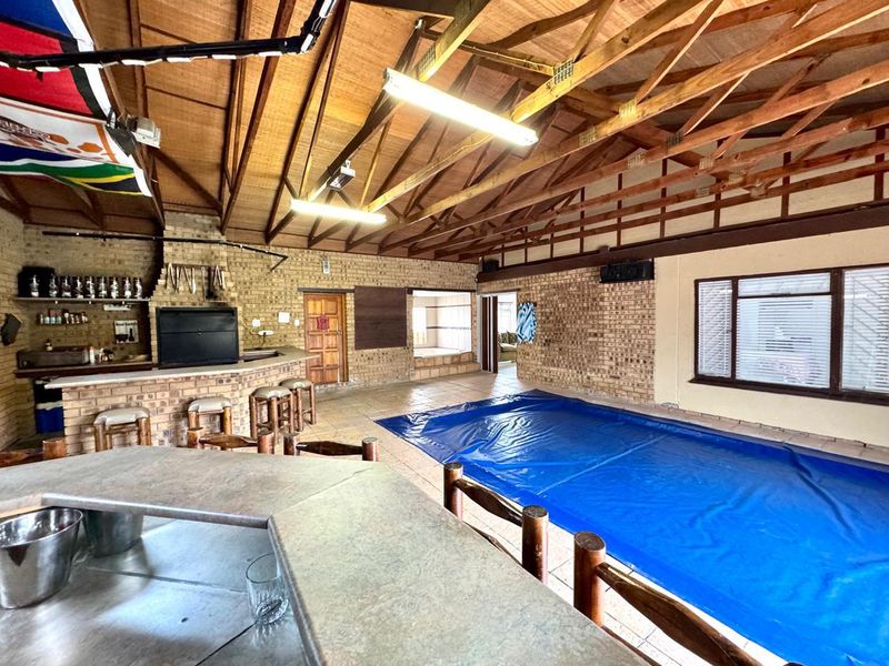 Spacious indoor entertainment area featuring a built-in bar, braai, and an integrated swimming pool under exposed timber roof trusses.