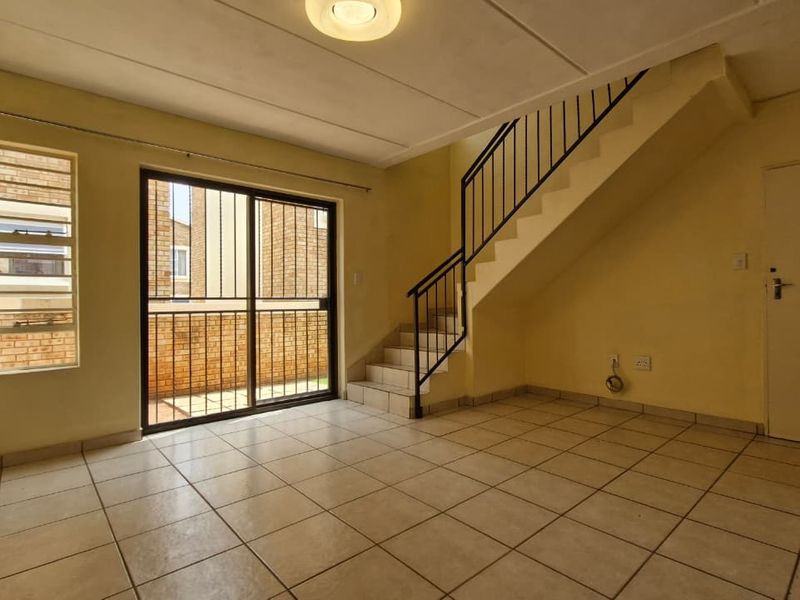 An empty, open-plan living area featuring tiled flooring, a staircase with a metal railing, and a sliding glass door leading to an outdoor space.