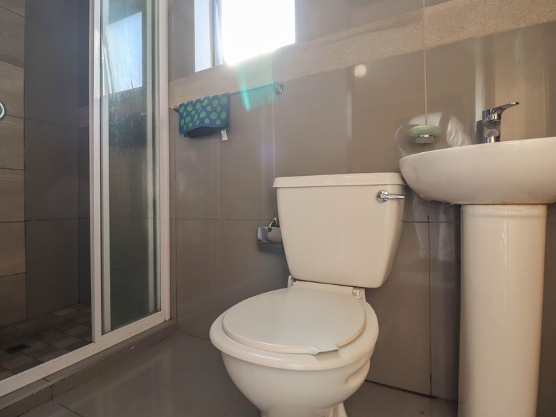A functional bathroom featuring a pedestal basin, a standard toilet, and a glass-enclosed shower with neutral wall and floor tiling.