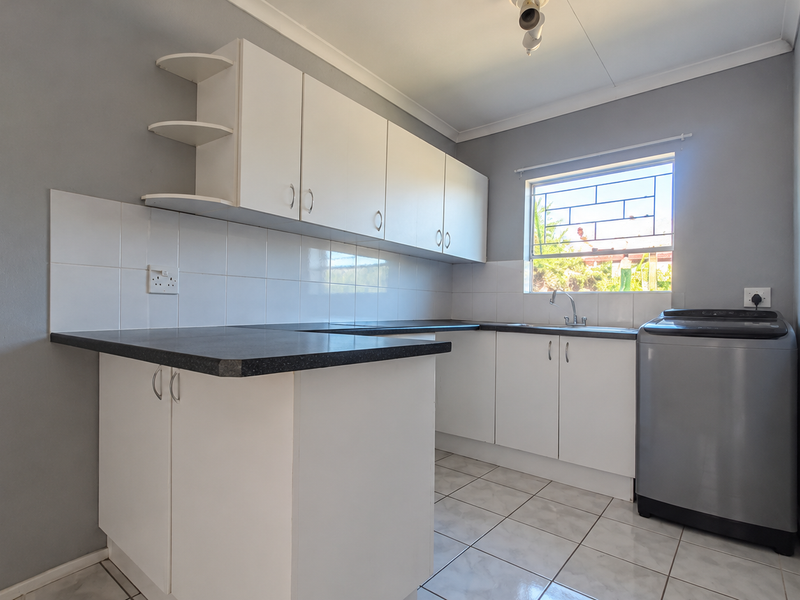 A functional kitchen featuring white built-in cabinetry, dark laminate countertops, tiled splashbacks, and a window with security bars.