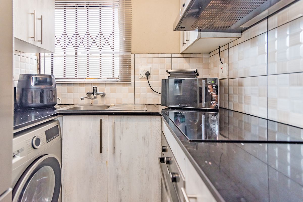 Compact kitchen featuring black granite countertops, built-in cabinetry, integrated hob, extractor fan, and a stainless steel sink with window security bars.
