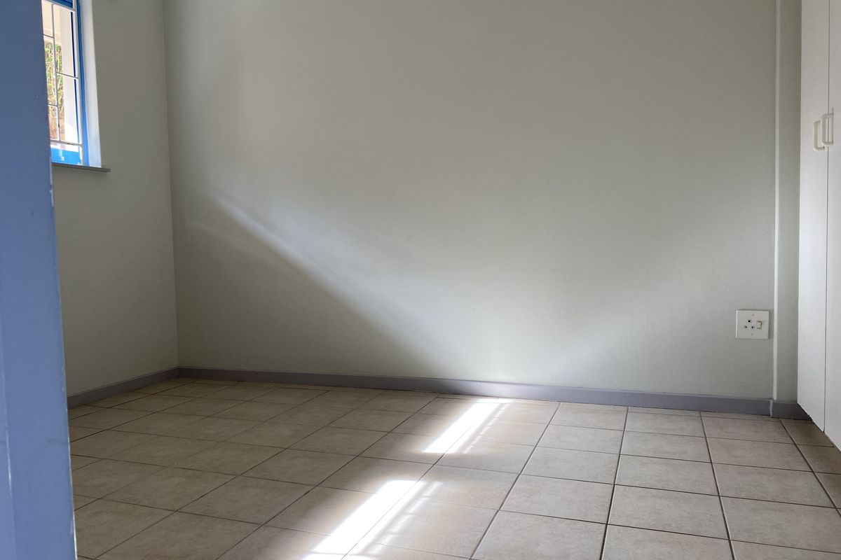 A bright, empty bedroom featuring neutral tiled flooring, a built-in cupboard, and a window with security bars.