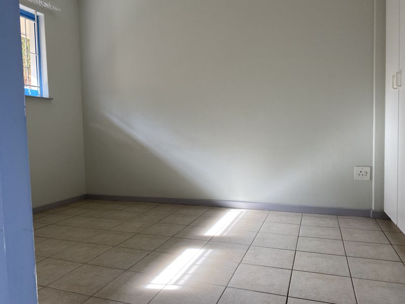 A bright, empty bedroom featuring neutral tiled flooring, a built-in cupboard, and a window with security bars.