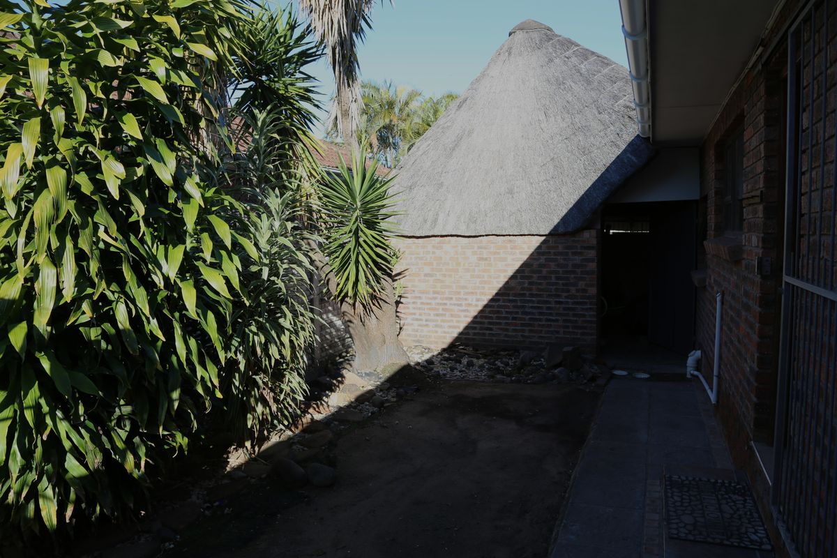 Rear exterior view featuring a traditional thatch roof structure, brick walling, and a paved walkway alongside an unlandscaped garden area.