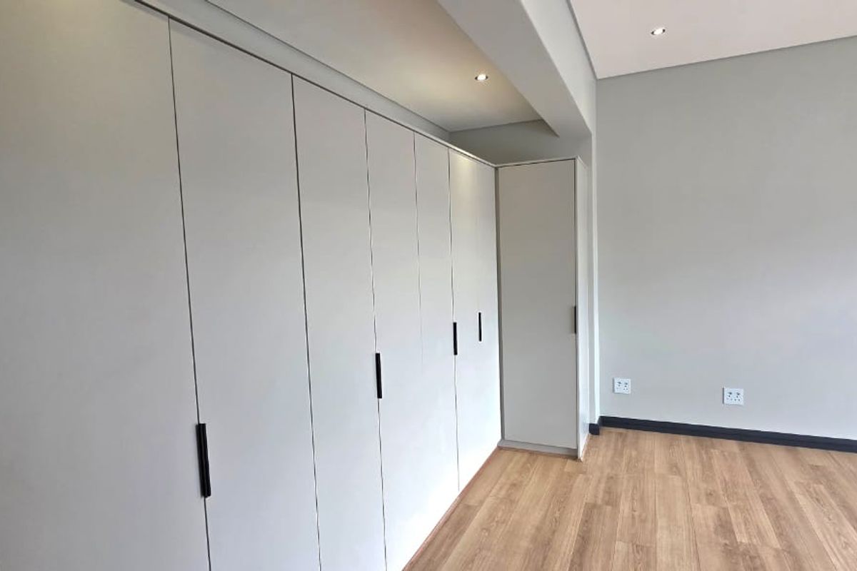 A bright, modern bedroom featuring extensive built-in cupboards, light laminate flooring, and clean, neutral walls with recessed lighting.
