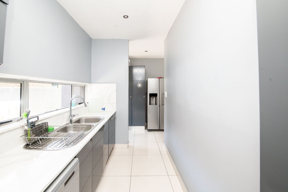A modern galley-style kitchen featuring sleek grey cabinetry, white countertops, a double stainless steel sink, and tiled flooring.