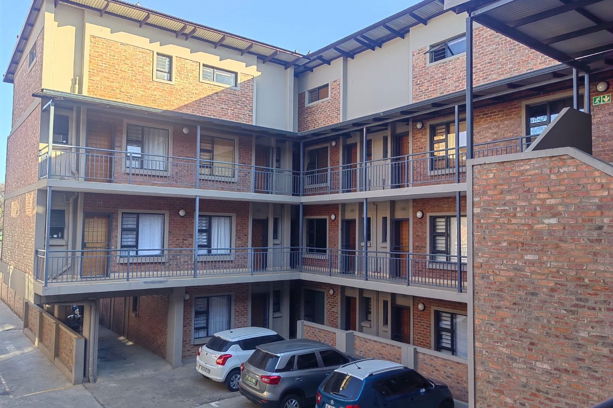 A multi-story apartment complex featuring face brick exterior walls, metal balcony railings, and a paved ground-level parking area.