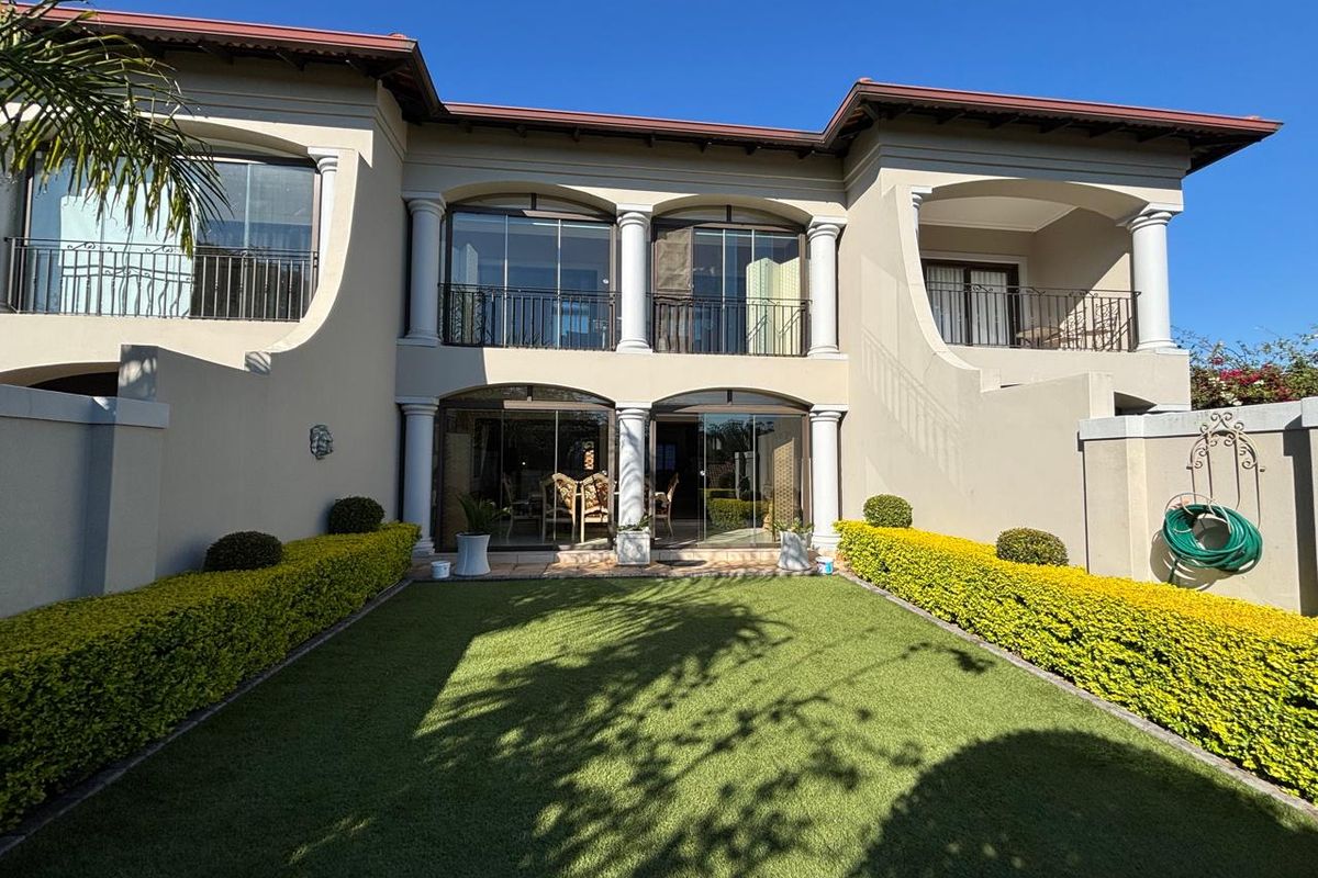 Rear view of a two-story townhouse featuring a manicured garden, artificial lawn, covered patio, and upper-level balconies with wrought iron railings.