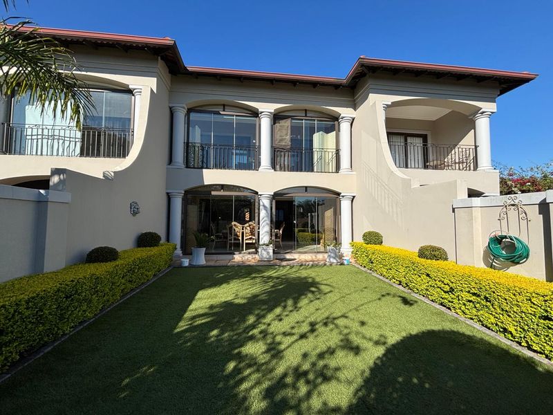 Rear view of a two-story townhouse featuring a manicured garden, artificial lawn, covered patio, and upper-level balconies with wrought iron railings.