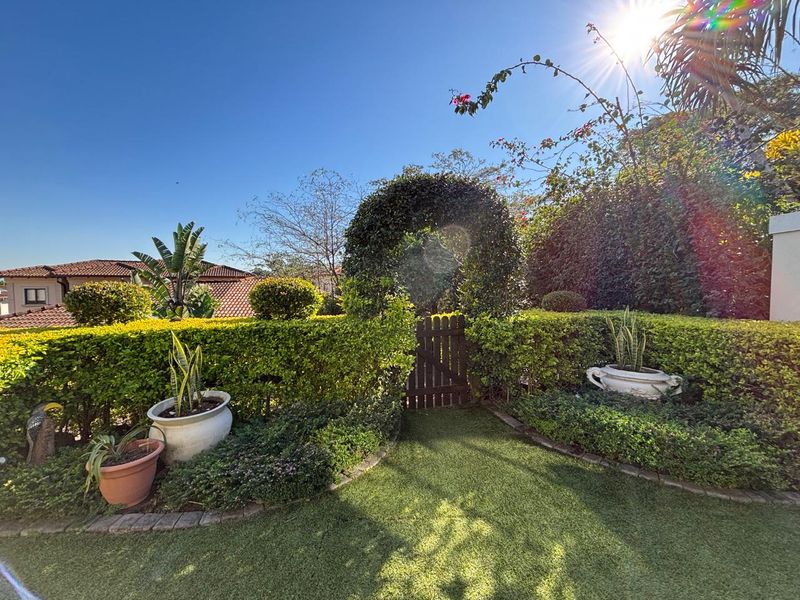 A well-maintained garden area featuring a manicured hedge, a charming wooden gate with an arched top, and neat artificial lawn with brick edging.