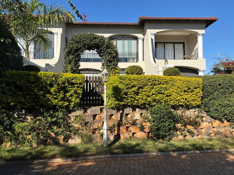 Front elevation of a two-story townhouse featuring a stone retaining wall, manicured hedges, and upper-level balconies with classical architectural columns.