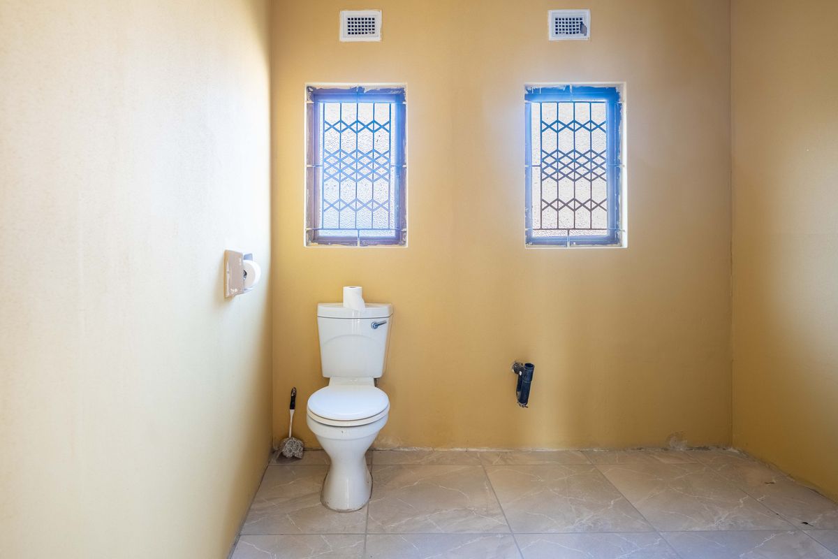 A basic bathroom featuring a toilet, tiled flooring, and security-barred windows, currently requiring finishing touches to the exposed plumbing and wall base.