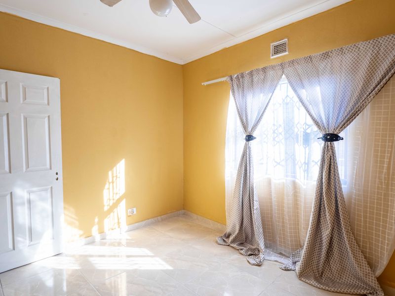 A bright, empty bedroom featuring tiled flooring, a ceiling fan, and a window fitted with a security gate.