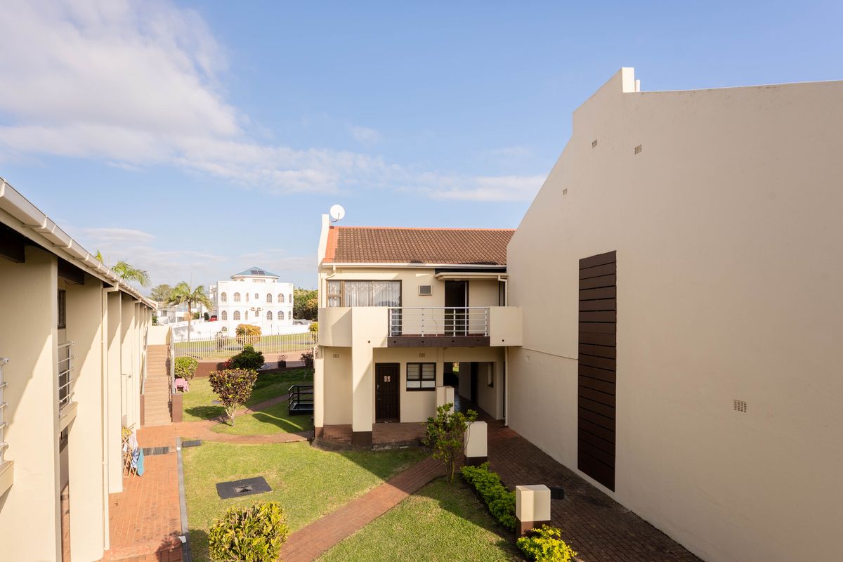 A view of the rear exterior of the flat complex, featuring a balcony, paved walkways, and well-maintained communal garden areas.