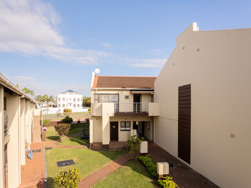 A view of the rear exterior of the flat complex, featuring a balcony, paved walkways, and well-maintained communal garden areas.