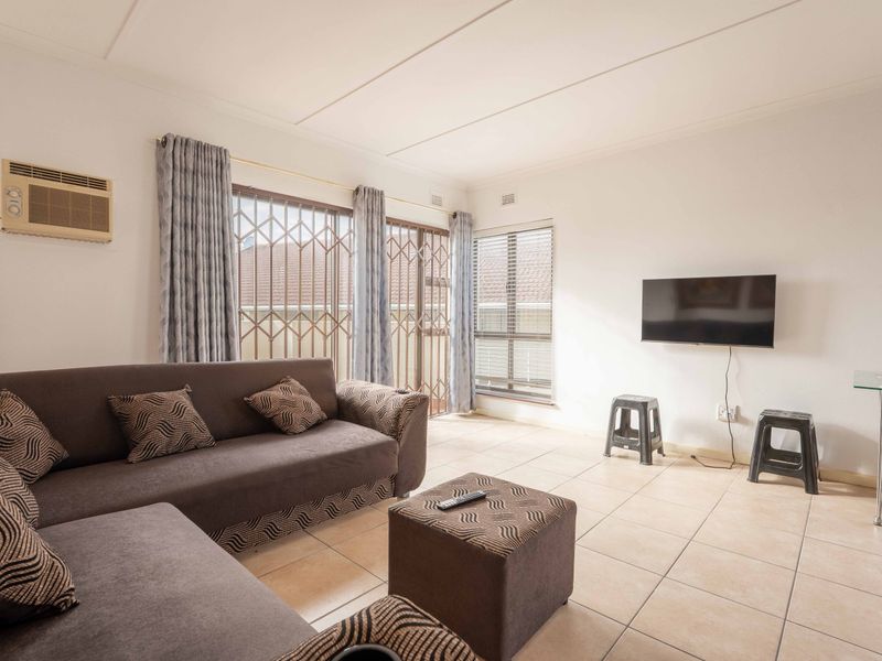 A bright living room featuring tiled flooring, a wall-mounted air conditioning unit, and secure window gates, offering a functional and well-lit living space.