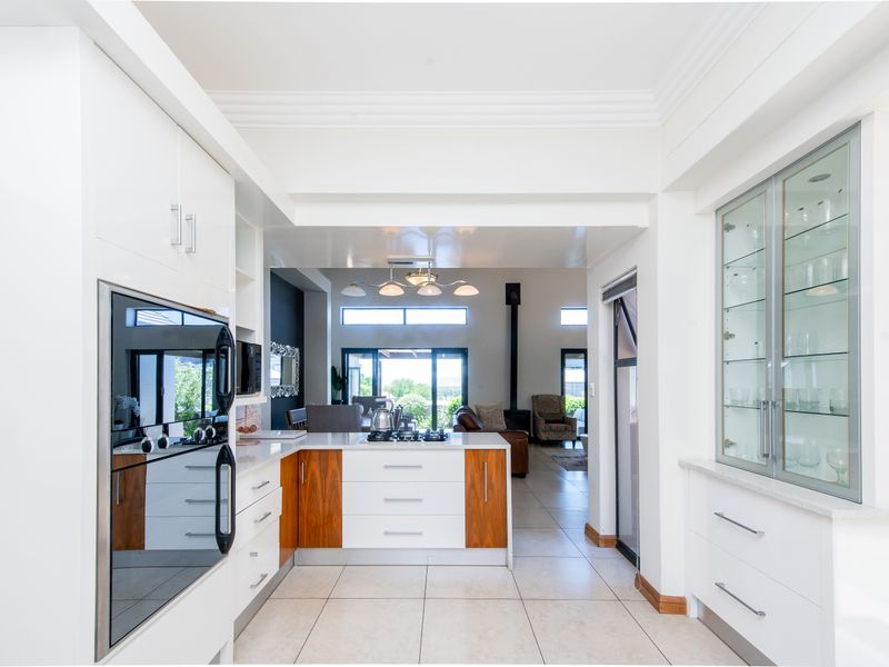 Modern kitchen featuring sleek white and wood-toned cabinetry, built-in oven, gas hob, and elegant glass-fronted display units, flowing into an open-plan living area.