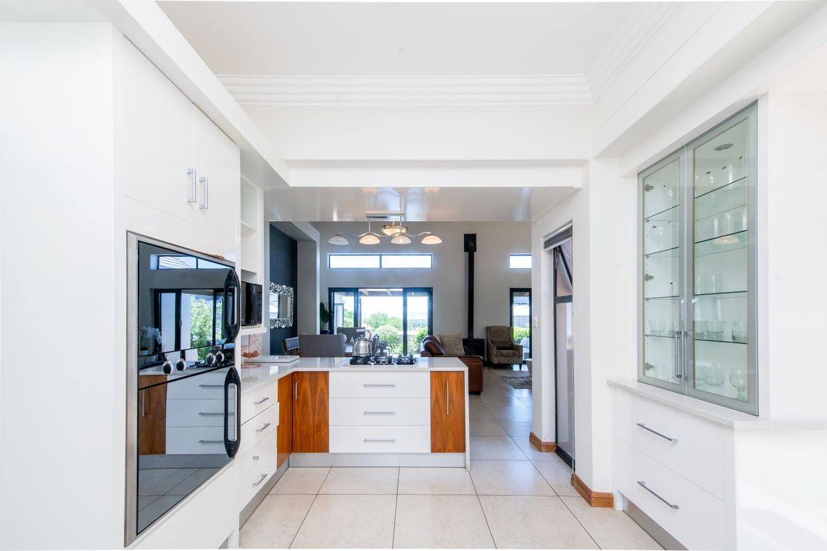 Modern kitchen featuring sleek white and wood-toned cabinetry, built-in oven, gas hob, and elegant glass-fronted display units, flowing into an open-plan living area.
