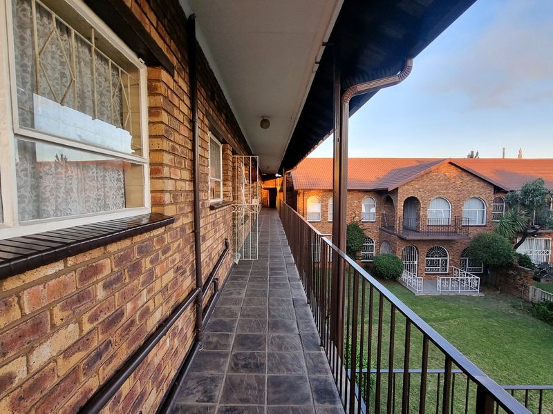 An elevated exterior walkway or balcony featuring brick walls, tiled flooring, and metal safety railings overlooking the complex grounds.