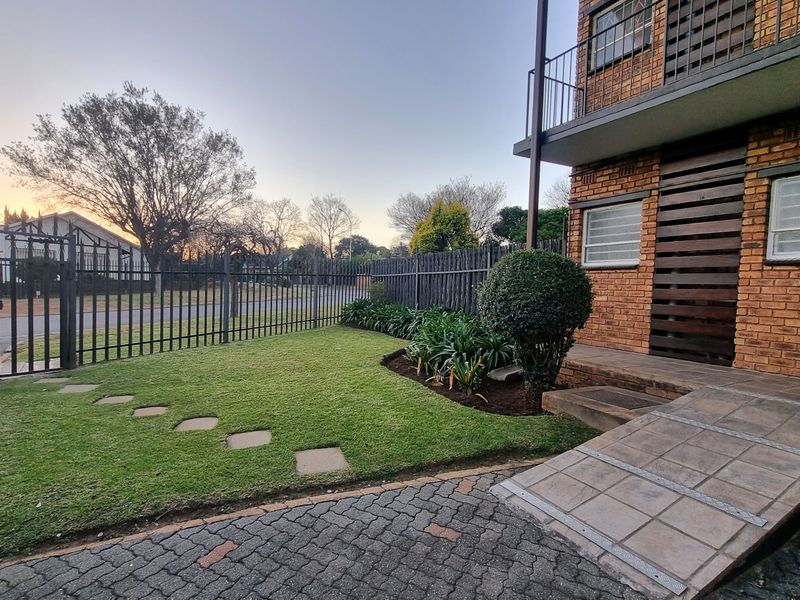 Front exterior view of a townhouse featuring a brick facade, paved driveway, accessible tiled entrance ramp, and a well-maintained garden with security fencing.