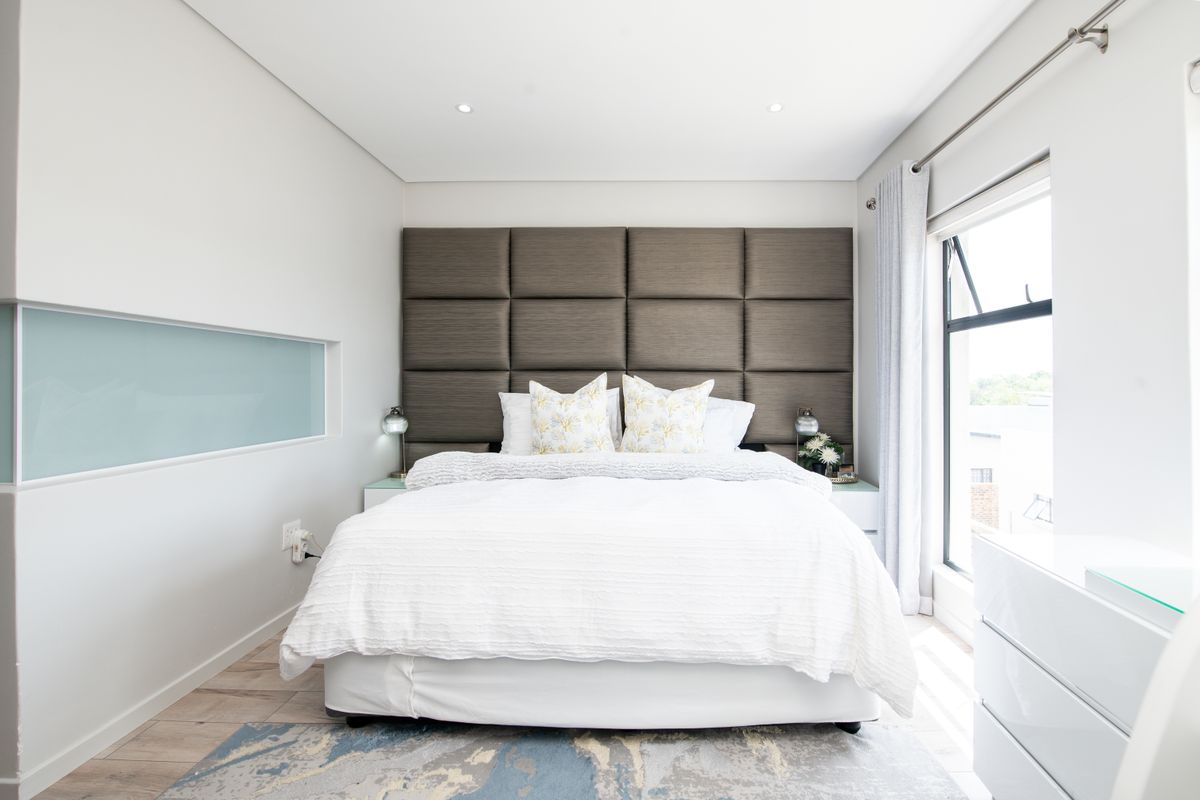 A modern, well-lit bedroom featuring a large upholstered headboard, recessed ceiling lighting, and a horizontal feature window.