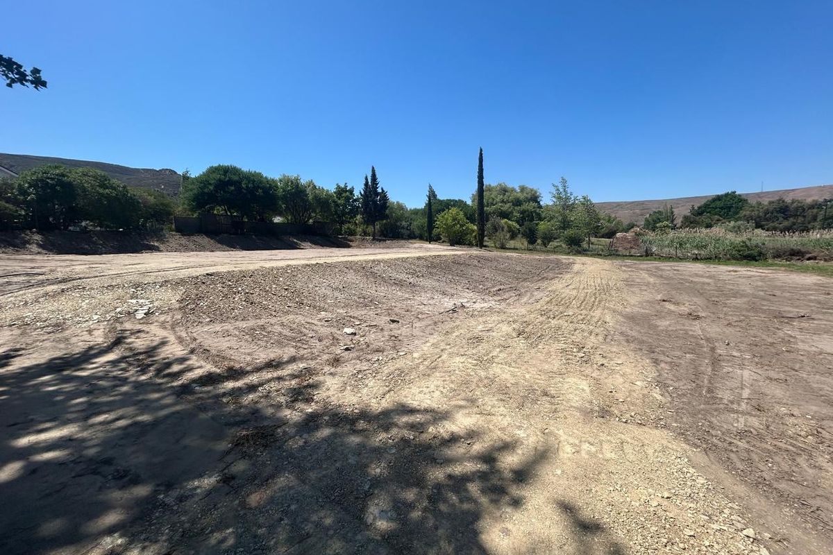 A wide-angle view of a vacant residential plot featuring cleared, level earth with surrounding mature trees and a clear blue sky.