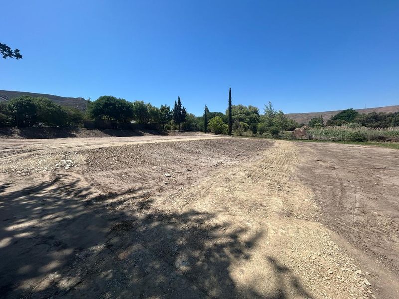 A wide-angle view of a vacant residential plot featuring cleared, level earth with surrounding mature trees and a clear blue sky.