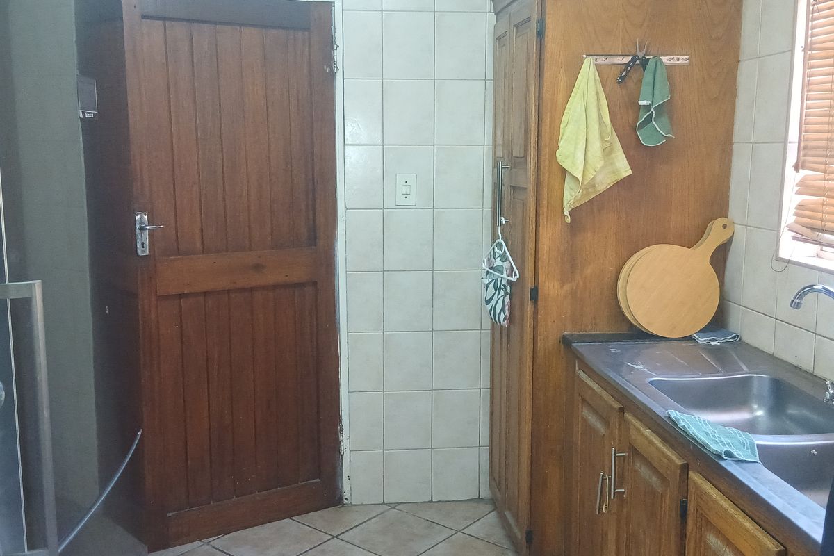 A functional kitchen area featuring wooden cabinetry, a stainless steel sink, tiled splashbacks, and tiled flooring with a wooden door entrance.