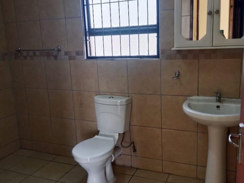 A functional bathroom featuring tiled walls and flooring, a pedestal basin, and a standard toilet, with a window fitted with security bars.