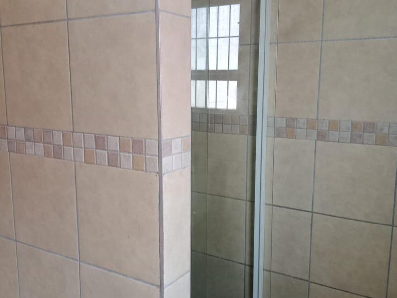 A functional bathroom shower area featuring tiled walls, a glass shower door, and a window with security bars.