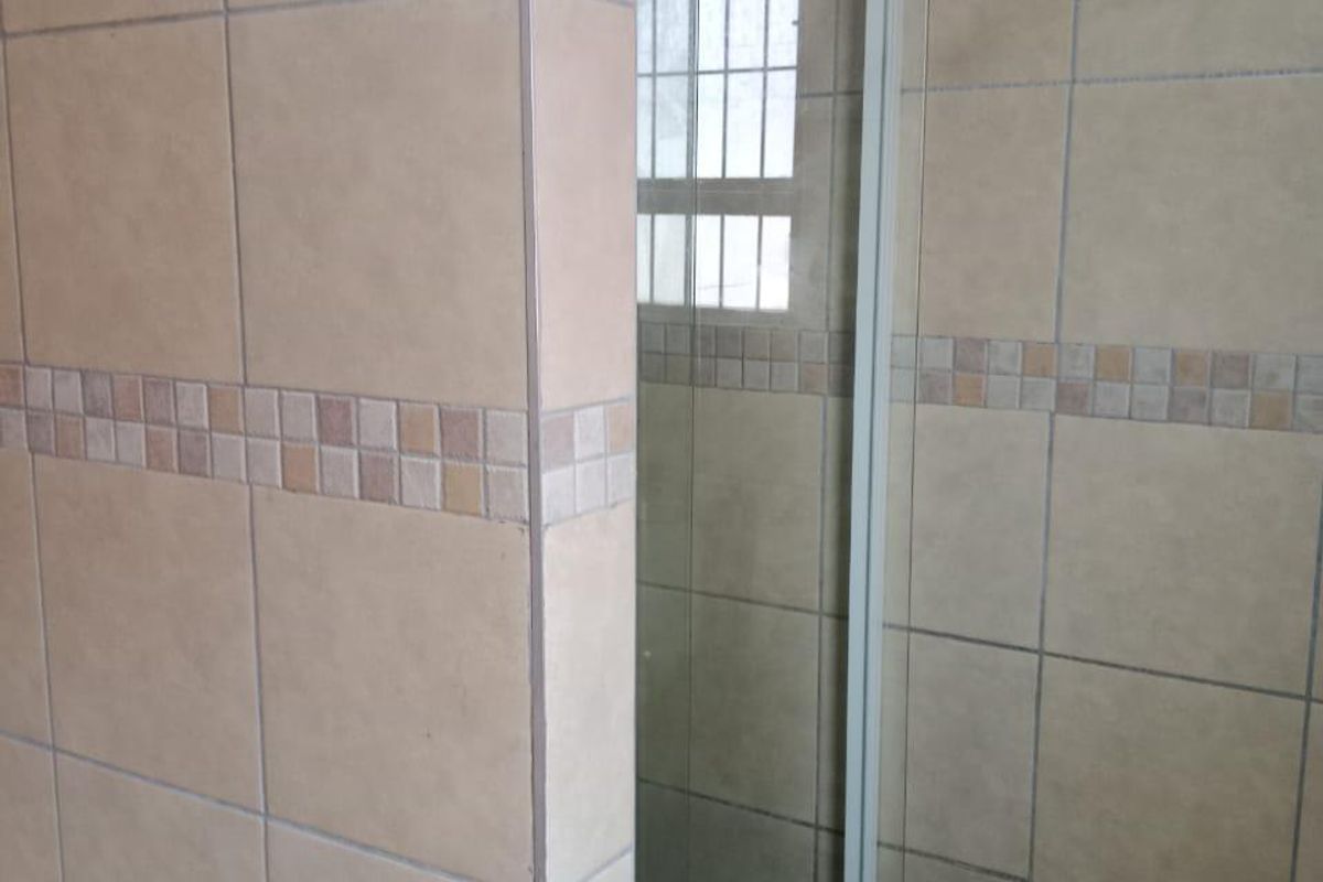 A functional bathroom shower area featuring tiled walls, a glass shower door, and a window with security bars.