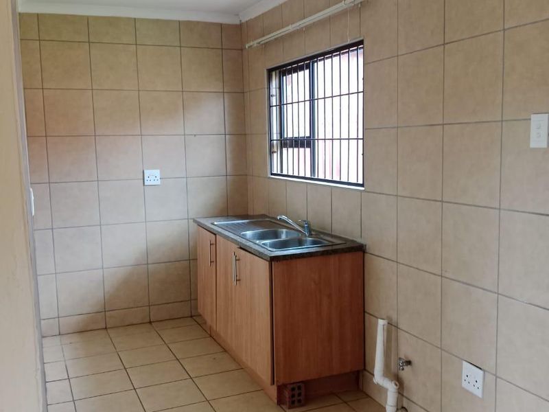 A basic, empty kitchen area featuring tiled walls and flooring, a single base cabinet with a sink, and security bars on the window.