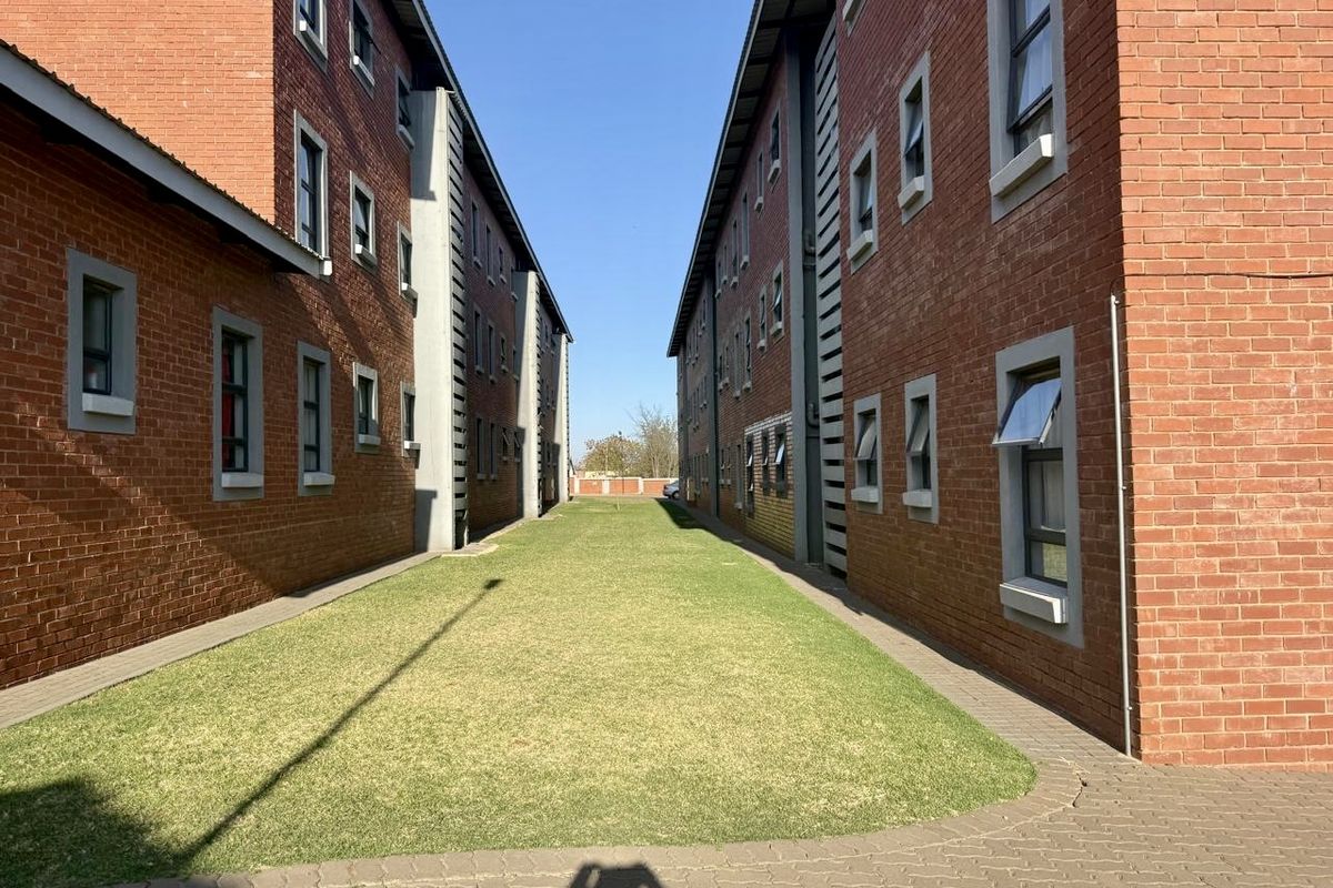 A view of the exterior space between two face brick apartment blocks, featuring a well-maintained lawn and paved pathways.