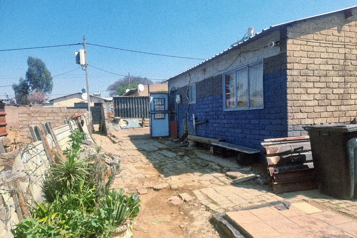 Rear exterior view of a brick house featuring a paved walkway, boundary wall, and basic utility infrastructure in a residential setting.