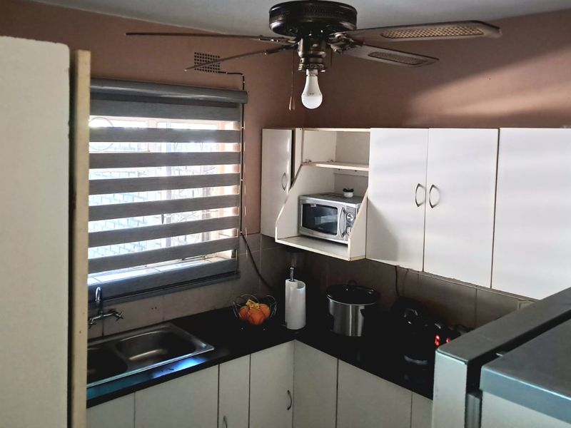 Compact kitchen featuring white built-in cabinetry, dark laminate countertops, a double basin sink, and a ceiling fan with integrated lighting.