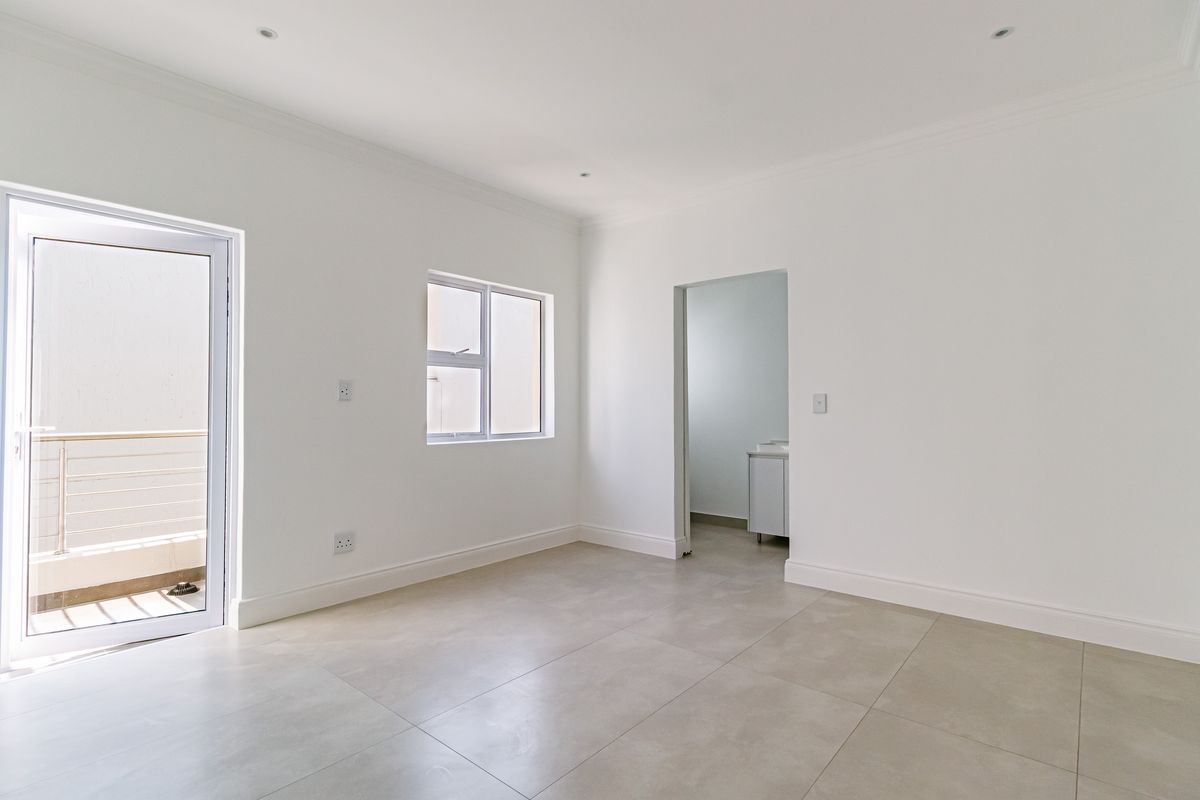 A bright, vacant bedroom featuring neutral tiled flooring, recessed lighting, and direct access to a private balcony through a glass door.