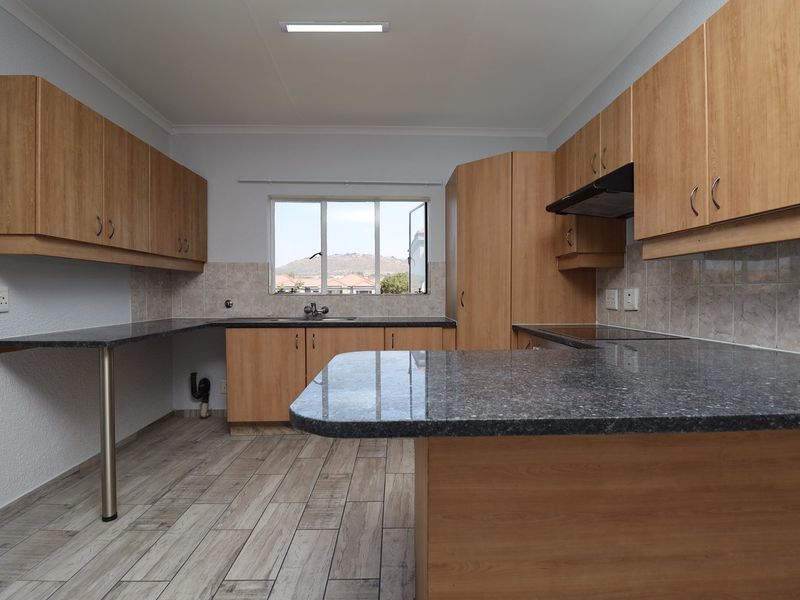 A clean, functional kitchen featuring wood-look cabinetry, granite countertops, built-in hob, extractor fan, and tiled flooring with a view through the window.