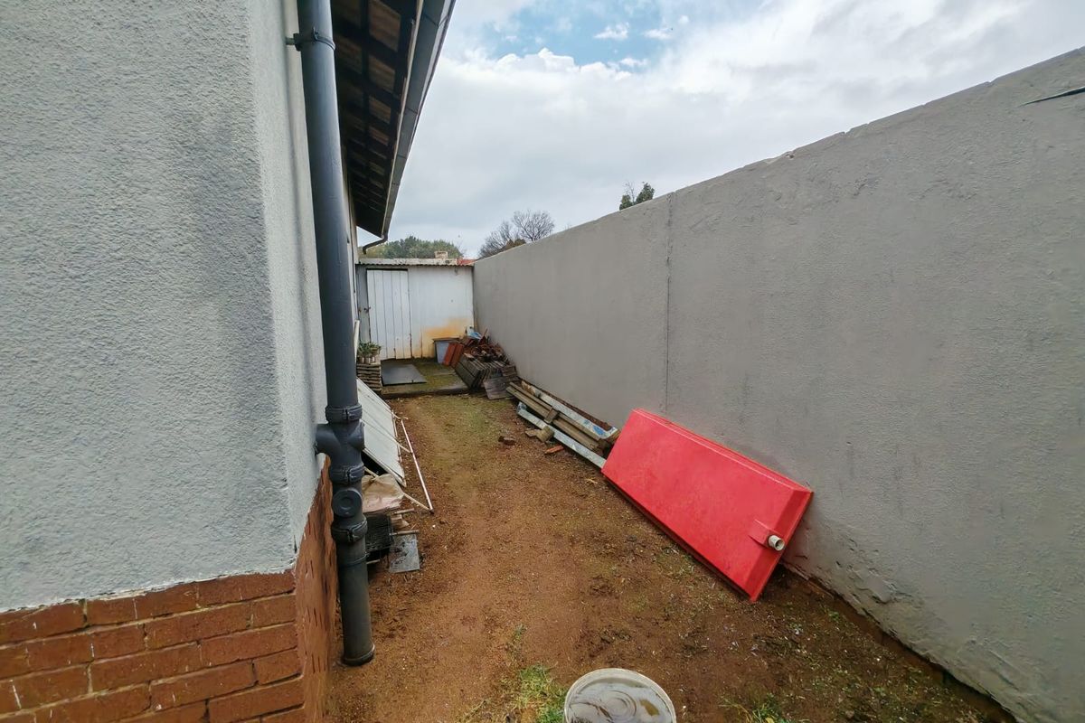 A narrow side exterior passage featuring a high boundary wall, exposed brickwork, and a downpipe, leading toward a small storage structure.