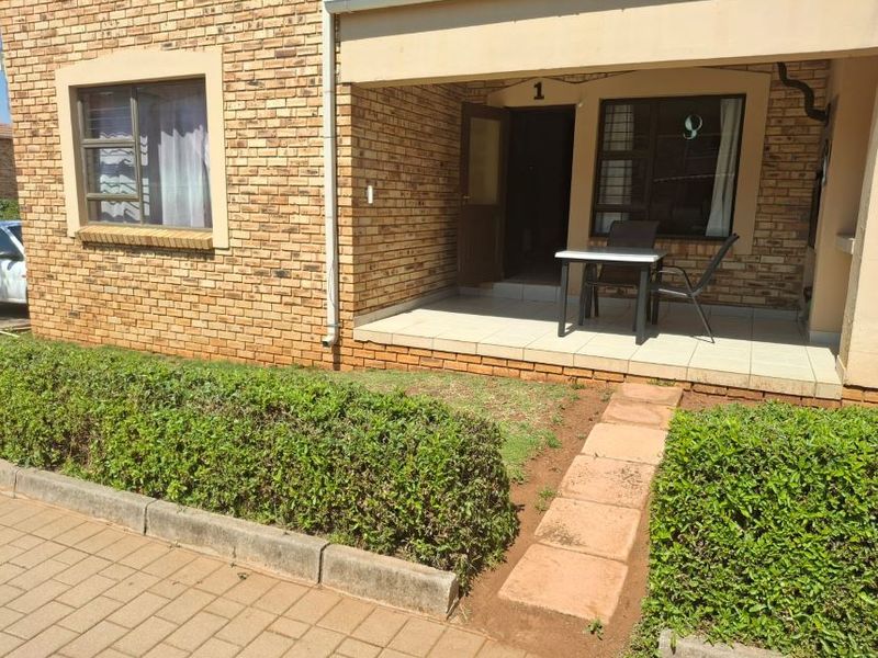 Front view of an apartment unit featuring a face brick facade, a covered tiled patio entrance, and a small landscaped garden with a paved walkway.