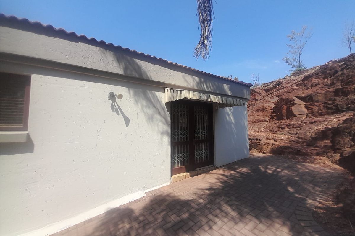 Rear exterior view of the property featuring a paved patio area, security gate access, and a protective awning against a rocky landscape backdrop.