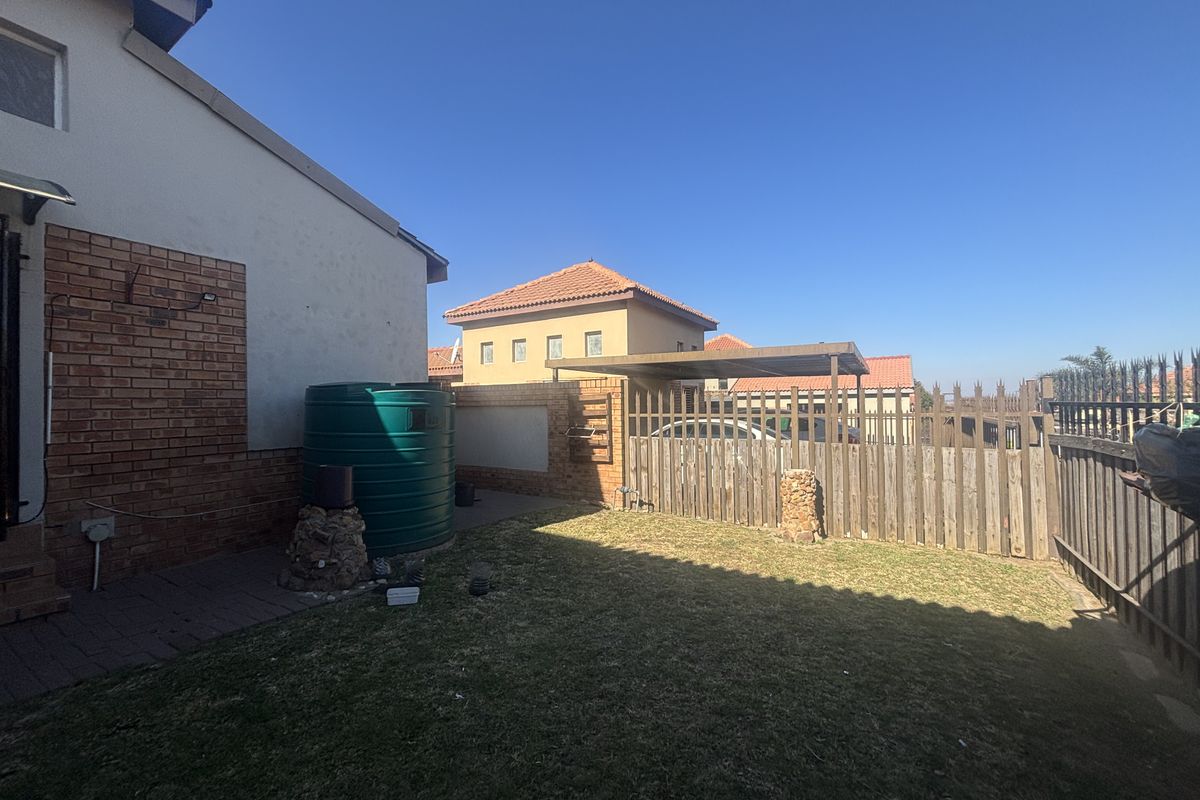 A rear exterior view of the townhouse featuring a small lawn, paved area, water tank, and timber boundary fencing with a carport.