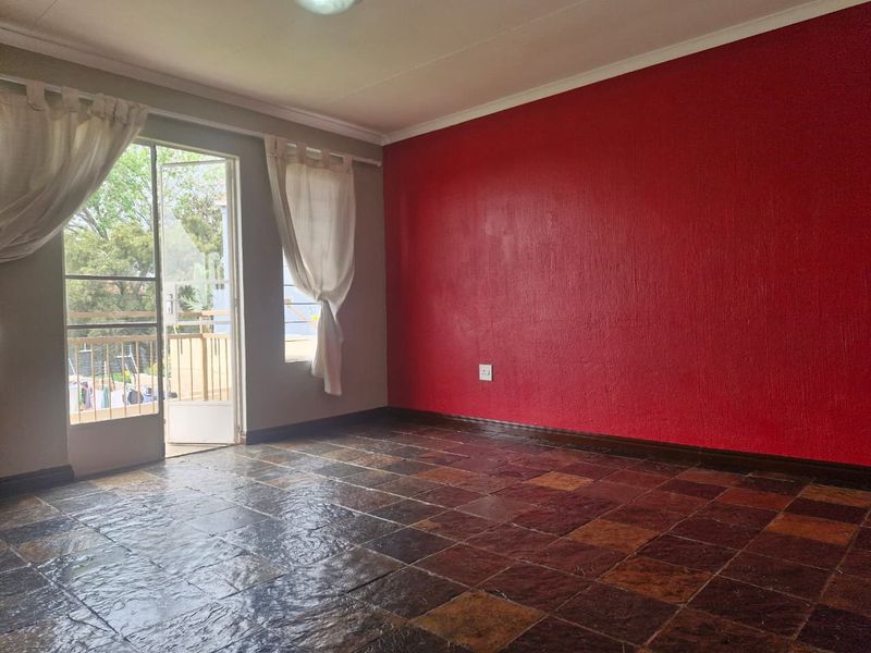 An empty living room featuring dark textured floor tiles, a bold red accent wall, and direct access to a balcony through a sliding glass door.