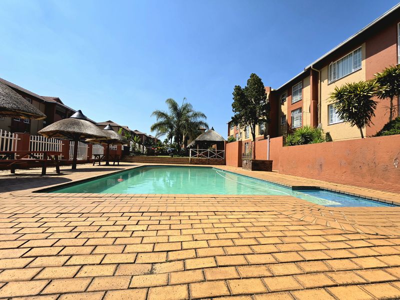 Communal swimming pool area featuring brick paving, thatched umbrellas, and surrounding residential townhouse blocks.