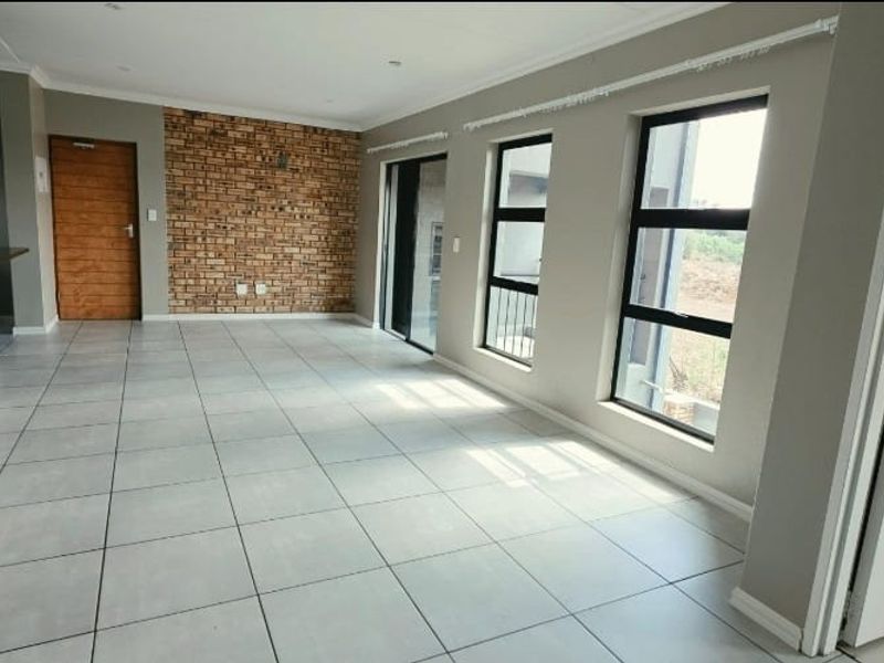 Spacious open-plan living area featuring modern tiled flooring, an exposed brick feature wall, and large windows providing ample natural light.