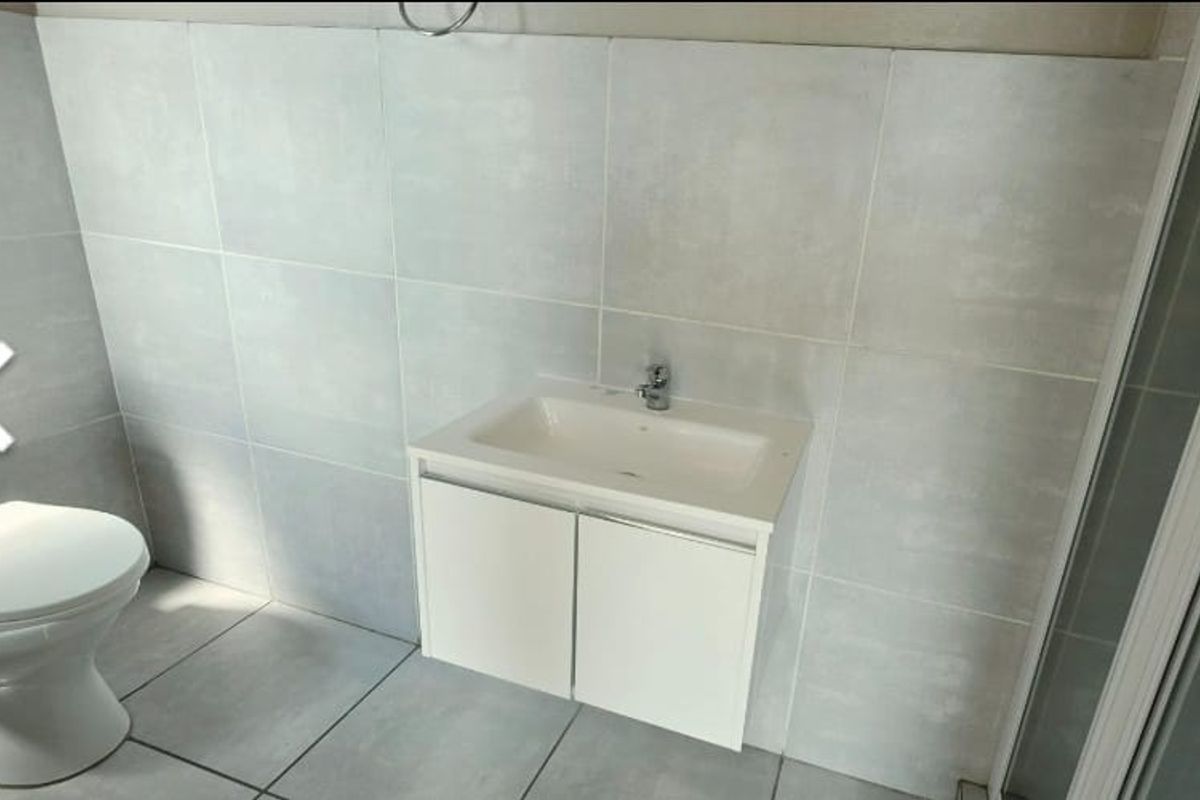 A clean, modern bathroom featuring a white vanity cabinet with integrated basin, a standard toilet, and a glass-enclosed shower with neutral wall tiling.