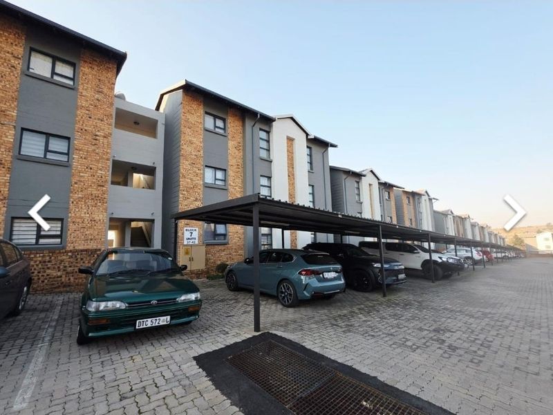 Exterior view of the apartment complex featuring a paved parking area with covered bays and modern brick and grey facade finishes.