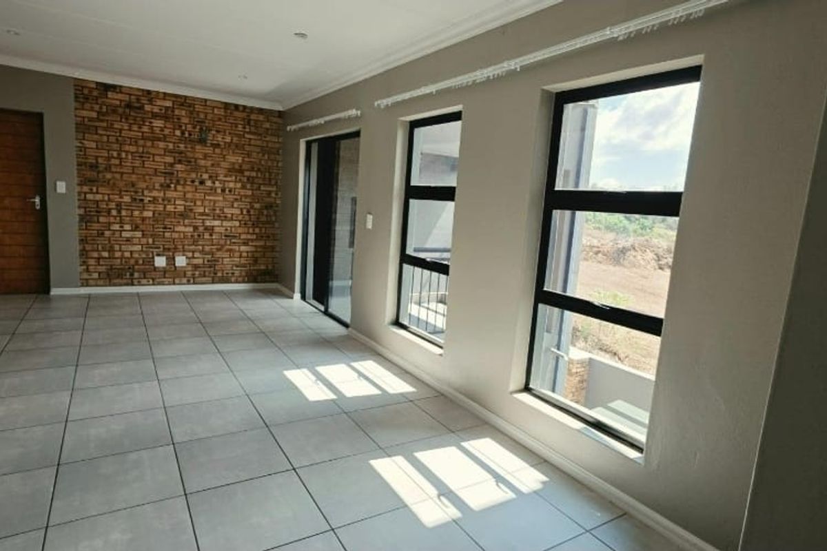 A bright, empty living area featuring modern grey floor tiles, an exposed brick feature wall, and large aluminium-framed windows providing natural light.